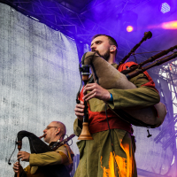 Two musicians perform on stage with bagpipes at what seems to be a festival. The man in the foreground, wearing a green robe and red sash, plays intently, while the man in the background also focuses on his playing. The stage is bathed in moody purple and blue lighting, with a large backdrop screen behind them.