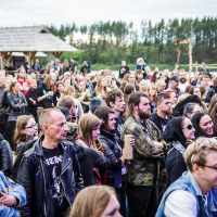 A large crowd of people, predominantly dressed in dark clothing and leather jackets, are gathered outdoors for Kilkim Žaibu 2015. The background features a wooded area with green trees and a rustic wooden shelter. Attendees appear to be enjoying the event under a cloudy sky.