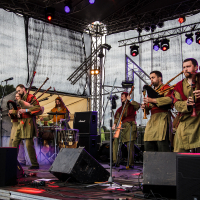 At the 2015 Kilkim Žaibu festival, a band of musicians dressed in earth-toned medieval-style clothing performs on an outdoor stage. Some play bagpipes while others beat drums. Colorful lights illuminate the scene, and various speakers and equipment are visible amidst a large netted backdrop surrounding the stage area.
