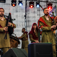 A group of musicians dressed in medieval-style tunics with red scarves play bagpipes and drums on a stage. The backdrop, reminiscent of the vibrant atmosphere at Kilkim Žaibu 2015, is lit with colorful lights, and musical equipment is visible in the foreground. A woman in the center appears to adjust an instrument.