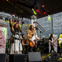 A band performs on stage under colorful lights at the 2015 Kilkim Žaibu outdoor event. The frontman, dressed in medieval-inspired clothing, raises his hand while another musician plays a guitar. Other band members are visible, and the backdrop features banners with artistic designs. Equipment and speakers are visible in the background.