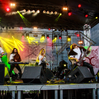 A live band performs on stage with dynamic lighting effects reminiscent of Kilkim Žaibu 2015. The lineup includes a keyboardist, a female vocalist, and three male guitarists. The stage is adorned with red and yellow lights, and a backdrop featuring the band's logo. The musicians are energetically engaged in their performance.
