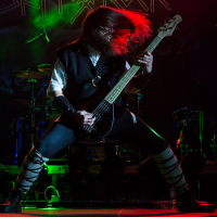 A musician with long hair thrashes their head while playing a black electric guitar on stage at Kilkim Žaibu 2015. They are wearing a dark outfit with light accents on the legs and arms. The background is dimly lit with green and purple lights, showcasing musical instruments and equipment.