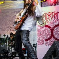 A musician with long hair plays an electric guitar onstage at the 2015 Kilkim Žaibu festival. He wears a white shirt and black leather pants with side lace detailing. In the background, a drummer plays a drum kit. The stage is decorated with ornate artwork, and colorful lights illuminate the scene.