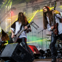 A rock band performs energetically on stage at the 2015 Kilkim Žaibu festival. The female singer points towards the crowd while two male guitarists, both with long hair, play intensely. Bright stage lights and colored beams create a dynamic atmosphere. A drummer is visible in the background. Band's name is partially visible on the backdrop.