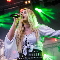 A woman with long blonde hair performs on stage at the Kilkim Žaibu festival. She holds a microphone, wearing a white blouse with flared sleeves and black pants adorned with a studded belt and laces. Green and purple stage lights illuminate the background, and her arm is raised as she sings passionately.