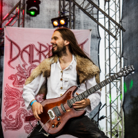 A musician with long hair, wearing a white shirt and fur vest, plays an electric guitar on stage at Kilkim Žaibu festival. Colorful stage lights illuminate him, and a banner with intricate red designs is in the background. He appears energetic and engaged with the performance, smiling broadly.