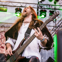 A musician with long hair and a beard passionately plays an electric bass guitar on stage at the 2015 Kilkim Žaibu festival. He is wearing a white shirt and black pants with a fur garment over his shoulders. Colorful stage lights and scaffolding are in the background, and the musician's mouth is wide open as if singing or shouting.