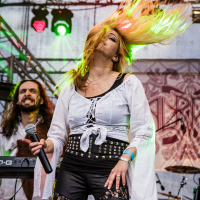 A woman on stage at the 2015 Kilkim Žaibu festival, with long hair flying mid-air, is energetically performing. She is wearing a white blouse, black pants with lace cutouts, and a studded belt. A man in the background is playing a keyboard. The stage is lit with red and green lights against an intricate backdrop.