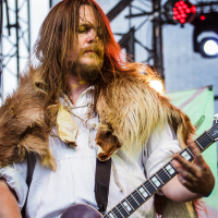 A musician with long hair performs on stage at Kilkim Žaibu 2015, playing an electric guitar. He is wearing a fur cloak over a white shirt. The stage has metal scaffolding and lights in the background. The atmosphere is vibrant, with a focus on his intense expression and dynamic posture.

Credit: xltphoto.net.