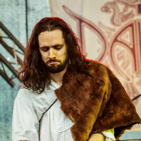 A musician with long, dark hair and a beard plays a Roland Juno-G synthesizer on stage at Kilkim Žaibu 2015. He wears a white shirt and a brown fur stole over one shoulder. The background features part of a large emblem or logo with intricate designs and the partial letters "DA".