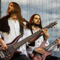 At the 2015 Kilkim Žaibu festival, two musicians with long hair, dressed in medieval-style white shirts and fur cloaks, are playing electric guitars on an outdoor stage. The foreground musician plays a bass guitar, while the background musician plays a rhythm guitar, both deeply focused on their performance.