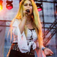A female singer with long blonde hair performs on stage at the 2015 Kilkim Žaibu festival, holding a microphone in her right hand. She wears a white blouse with laces at the front, black pants with sheer panels, and a studded black belt. The background features stage lights and rigging. Photo credit "xltphoto.net" is at the bottom.