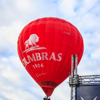 A red hot air balloon with the logo of "Stumbras 1906" and a white bison graphic rises against a blue sky with scattered clouds at the Kilkim Žaibu 2015 festival. The balloon's identification "LY-OMO" is visible. In the foreground is a metal scaffolding structure with a partial logo at the bottom right.