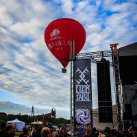 A bright red hot air balloon with white text floats above a crowd at the 2015 Kilkim Žaibu festival. Banners on the right bear logos and festival names. People on the ground observe the balloon amidst tents and structures under a partly cloudy blue sky. A mechanical dragon sculpture is visible in the background.