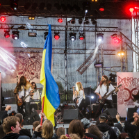 A band performs energetically on stage at an outdoor festival, with a large Ukrainian flag waved by an audience member in the foreground. The stage backdrop features the band's logo with "Kilkim Žaibu 2015" subtly included. The crowd is engaged, some raising their hands, all amid vibrant and intense lighting.