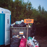 A large outdoor garbage bin at the 2015 Kilkim Žaibu festival is overflowing with trash, surrounded by more bags on the ground. A makeshift "VIP" sign humorously adorns the bin, while a portable toilet peeks out on the left. Tall trees provide a natural backdrop to this quirky festival scene.