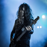 At the 2015 Kilkim Žaibu festival, a musician with long, wavy hair performs on stage. Their hair obscures their face as they play a bass guitar. The background is filled with blue and black lighting, creating a dramatic and intense atmosphere, while their dark outfit enhances the moody setting.