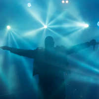A silhouetted figure stands with outstretched arms amidst a blue, hazy environment. Bright stage lights beam from behind, illuminating the background with starburst effects. The person, holding a microphone, adds an ethereal and dramatic touch reminiscent of Kilkim Žaibu festival 2015.