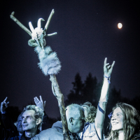 A crowd at the 2015 Kilkim Žaibu festival, with a full moon in the background. People are raising their hands, some making rock and roll signs. A prominently raised wooden staff adorned with feathers, fur, and a skull stands out in the crowd. The atmosphere is energetic and festive.