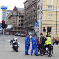 A street scene from 2010 with three people in blue uniforms and one in a yellow vest standing next to two bicycles. A police motorcycle is parked nearby. They're conversing on a city sidewalk amidst ornate historic buildings, possibly during a Kritiska masa event, with a traffic light and distant pedestrians in the background.