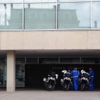 Three police officers, dressed in blue uniforms, are standing beside three police motorcycles under a concrete overhang. The building behind them features large windows on the upper floor and the words "DAILES TEATRIS" on the dark wall beneath the overhang. This scene evokes memories of Kritiska masa 2010.