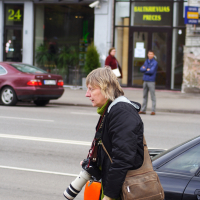 A person with short, light hair is walking on a city sidewalk, carrying a camera with a large lens. They are wearing a dark jacket and an olive scarf, and have a brown shoulder bag. In the background, cars are parked along the street, and people are standing in front of shopfronts during Kritiska masa 2010.