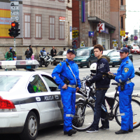 Two police officers in blue uniforms stand near a police car and a young man with a bicycle on a city street, reminiscent of the cycling event Kritiska masa in 2010. The conversation appears casual. Behind them, the cityscape features buildings, traffic lights, and pedestrians, suggesting routine urban activity.