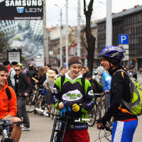 A group of people are gathered in a bustling city street with bicycles, reminiscent of Kritiska masa events around 2010. Three men in the foreground, smiling and chatting in casual and sporty attire with helmets and backpacks, stand out. The background features tall buildings, a "Samsung" advertisement, and more cyclists.