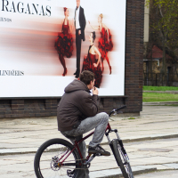 A person in a brown jacket and gray jeans is sitting on a bicycle on a paved walkway, reminiscent of the 2010 Kritiska masa. They appear to be eating an ice cream cone. Behind them, there is a large advertisement featuring a man in formal attire and women in red and black flamenco dresses. The sign has text in a non-English language.