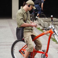 A young man in khaki shorts and a matching shirt sits on a bright orange bicycle. He wears a black cap with headphones, and a black shoulder bag across his body while looking at a smartphone. In the background, another cyclist with a blue helmet rides by during the 2010 Kritiska masa event in an outdoor urban area.