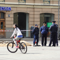 A woman rides a bicycle past a group of four police officers standing beside a red and white caution tape. They appear to be securing a construction area marked by the tape and some soil on the ground. The scene, reminiscent of Kritiska masa, unfolds in front of a beige building with green and blue trim around the windows.