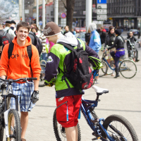 In an urban area during a 2010 Kritiska masa event, two young men stand with their bicycles. One wears a bright orange hoodie and plaid shorts, while the other dons a green jacket, red shorts, and a brown beanie. They converse cheerfully among a crowd of cyclists, with buildings and parked bikes visible in the background.