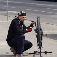 A person wearing a bandana, gloves, and a black sweatshirt is kneeling and working on an upside-down bicycle on a sidewalk during the Kritiska masa event in 2010. The background shows an unfocused street with a white and blue bus. The individual appears focused on adjusting the bike's pedal or chain.