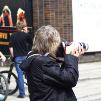 A person with gray hair, wearing a dark coat, photographs something with a large Canon camera attached to a telephoto lens. In the background of this 2010 scene, two young people with bikes are visible—one standing and one sitting—alongside a large poster and part of a building facade, perhaps hinting at Kritiska masa.