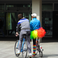 Two bicyclists in front of a building during the 2010 Kritiska masa event. The person on the left is wearing a gray outfit with blue stripes, while the person on the right is dressed in colorful clothing, including a blue jacket, neon tutu, and rainbow-striped leg warmers. Both are looking forward, their backs facing the camera.