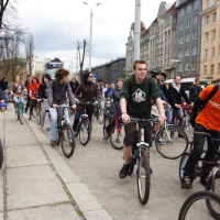 A large group of people of various ages is participating in a Kritiska masa bike ride along a city street. They are riding bicycles and appear to be in good spirits, wearing casual clothing. The backdrop features tall, colorful buildings and a partially cloudy sky, capturing the essence of this 2010 community event.