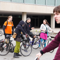 A group of five young adults stands together with their bicycles in an urban area, engaging in conversation. One person in focus rides a bike in the foreground. The background features a large building with glass windows, recalling the 2010 Kritiska masa vibes. The weather appears overcast, and everyone is dressed casually.