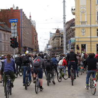A large group of cyclists rides down a city street bordered by both modern and historic buildings. The street, equipped with traffic lights and street signs, shows an overcast sky above. The cyclists, some wearing helmets, travel closely together in what looks like a Kritiska masa 2010 community ride or event.