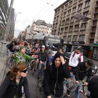 A large group of cyclists in casual attire ride along a city street, surrounded by tall, old buildings. Some cyclists wear helmets while others do not. The atmosphere appears busy and urban, reminiscent of the 2010 Kritiska Masa event, with a mix of buses and other structures in the background. The cyclists seem focused on their travel.