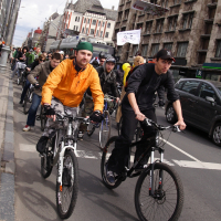 A group of cyclists is riding on a city street, reminiscent of the Kritiska masa event in 2010. Two men in the forefront are wearing an orange jacket and a black jacket, respectively. Behind them, more cyclists follow, with buildings lining both sides of the street. Some pedestrians can also be seen on the sidewalk to the left.