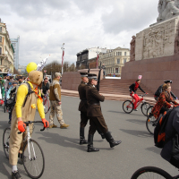 A group of people participates in a quirky cycling event, Kritiska masa 2010. Cyclists in colorful and eccentric costumes ride near a large monument. Two men in military uniforms stand in the center, one saluting. Balloons and flags add to the festive atmosphere against a backdrop of historical buildings.