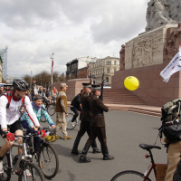 A busy urban scene in 2010 features a mix of cyclists, pedestrians, and onlookers in front of a large monument. People are casually dressed, some are pushing strollers, and one person is holding a yellow balloon. The sky is overcast, and buildings can be seen in the background, reminiscent of Kritiska masa events.