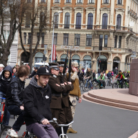 A group of cyclists from the Kritiska masa rides past a military ceremony in front of an ornate, historic building. Soldiers in various uniforms, including camouflage, are standing at attention while holding rifles. Spectators and additional cyclists are visible in the background. The sky is overcast, reminiscent of events from 2010.