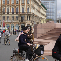 People ride bicycles on a city street near a grand historic building with ornate architecture and large windows. In the foreground, a person wearing a black coat and a beret cycles past, reminiscent of the 2010 Kritiska masa events, while others ride bikes in the background. A modern glass skyscraper is visible in the distance.