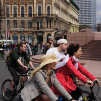 A group of people enjoy a bike ride through a city street during the 2010 Kritiska masa event. The joyful riders feature a man in a green hoodie, another in a pirate costume, a woman in a red polka-dot dress, and a woman in a beige hat and jacket. In the background are historic and modern buildings.