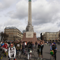 A large group of cyclists gathers in front of the Freedom Monument in Riga, Latvia for Kritiska masa 2010. The monument is tall with a statue of a woman holding three stars at the top. The cyclists, wearing casual clothing and helmets, are chatting and preparing to ride. Historic buildings and a cloudy sky are in the background.