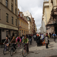 A bustling European street with historic buildings on both sides. Numerous pedestrians and cyclists navigate the cobblestone pavement, reminiscent of Kritiska masa 2010. A café named "Kafejnica ESPRESSO RIGA" is visible on the left. Various stalls and umbrellas line the right side, indicating a street market. The sky is overcast.