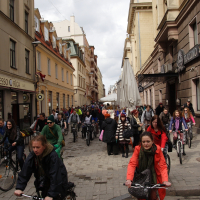 A busy, narrow street in a European city is filled with people riding bicycles and walking. The street, reminiscent of the 2010 Kritiska masa movement, is lined with tall historic buildings mixed with modern storefronts. Pedestrians and cyclists in vibrant clothing move actively under a cloudy sky, creating a lively atmosphere.