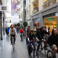 A group of people is riding bicycles through a modern, indoor shopping arcade with a glass ceiling, reminiscent of the 2010 Kritiska masa event. They are passing various stores, including a clothing store and a grocery store. Shoppers and pedestrians are visible in the background while a large advertisement banner hangs from the ceiling.
